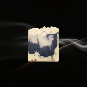 Handcrafted cold‑pressed soap bar with a natural beige base, speckled texture, and a blue‑and‑white artistic inlay resembling a bird, displayed against a dark background with soft smoke for a dramatic, premium presentation.