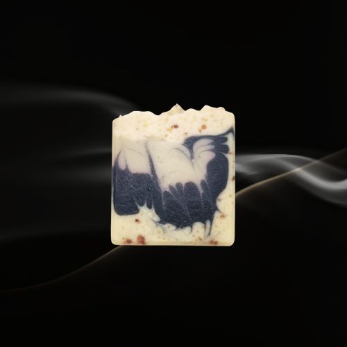 Handcrafted cold‑pressed soap bar with a natural beige base, speckled texture, and a blue‑and‑white artistic inlay resembling a bird, displayed against a dark background with soft smoke for a dramatic, premium presentation.