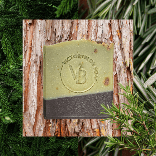 VICTORYBLOKS Rustic Evergreen soap bar with a green and brown split design, lightly speckled with Black African Soap Chips, set in front of fir branch, brown clay chips, and natural cedarwood. Made with shea butter, coconut oil, rice bran oil, clays, essential oils, and colloidal oat for a fresh, family-friendly cleanse.
